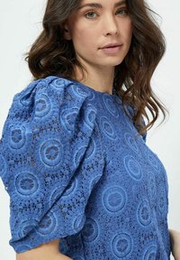 Blusa blu ricamata con maniche a sbuffo, che presenta motivi in pizzo circolari e tessuto strutturato. La vista ravvicinata mette in risalto i dettagli intricati.