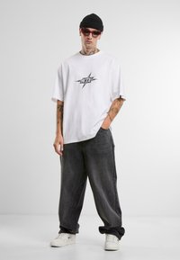 Hvid oversized t-shirt med et centralt sort grafisk tryk. Mørkegrå baggy jeans. Hvide sneakers og en sort strikbeanie. Lyserøde solbriller.
