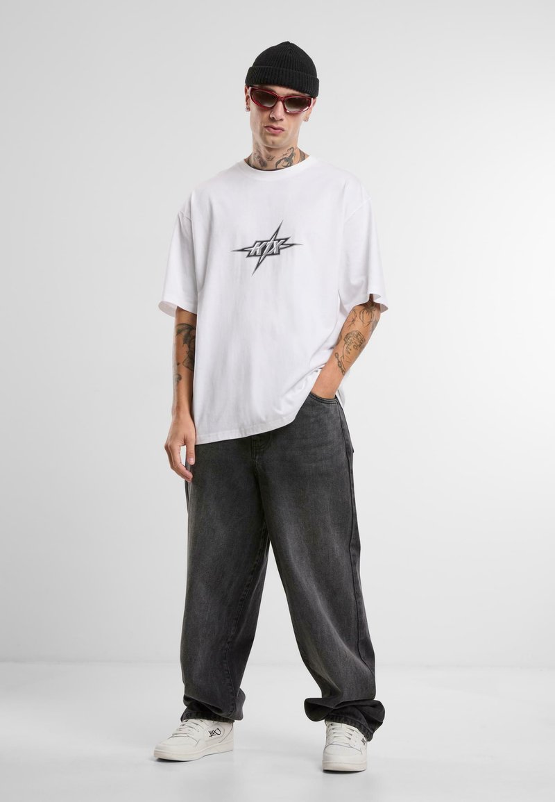 Hvid oversized t-shirt med et centralt sort grafisk tryk. Mørkegrå baggy jeans. Hvide sneakers og en sort strikbeanie. Lyserøde solbriller.
