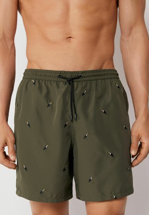 Man wearing olive green swim shorts with elastic waist and black drawstring, decorated with small toucan bird prints.