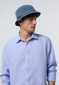 Cappello a secchiello blu con tessuto texturizzato e logo "NORTH SAILS" arancione. Camicia azzurrina con colletto classico e chiusura a bottoni.