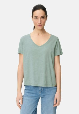 Kvinde med mørkt hår bundet tilbage iført lysegrøn V-udskæring kortærmet skjorte og blå jeans, set forfra mod en ensfarvet baggrund.