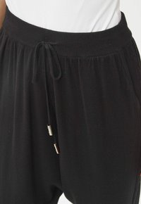 Joggers negros con una cintura acanalada, que cuentan con un cordón con puntas de metal plateado. Textura suave y corte relajado para mayor comodidad.