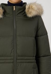 Olīvu zaļš pūkainsjakas ar augstu apkakli, kurai ir noņemama gaiši brūna mākslīgās kažokādas kapuce un priekša ar rāvējslēdzēju, izcelta ar pāršūtu polsterējumu.