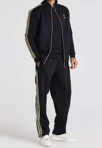 Chándal navy que incluye una chaqueta con cremallera y un escudo bordado, rayas doradas en las mangas y los pantalones, y zapatillas blancas.