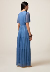 Rochie maxi albastră, cu volane scurte, realizată dintr-un material texturat cu sclipici și o talie ajustată, accesorizată cu brățări maro.