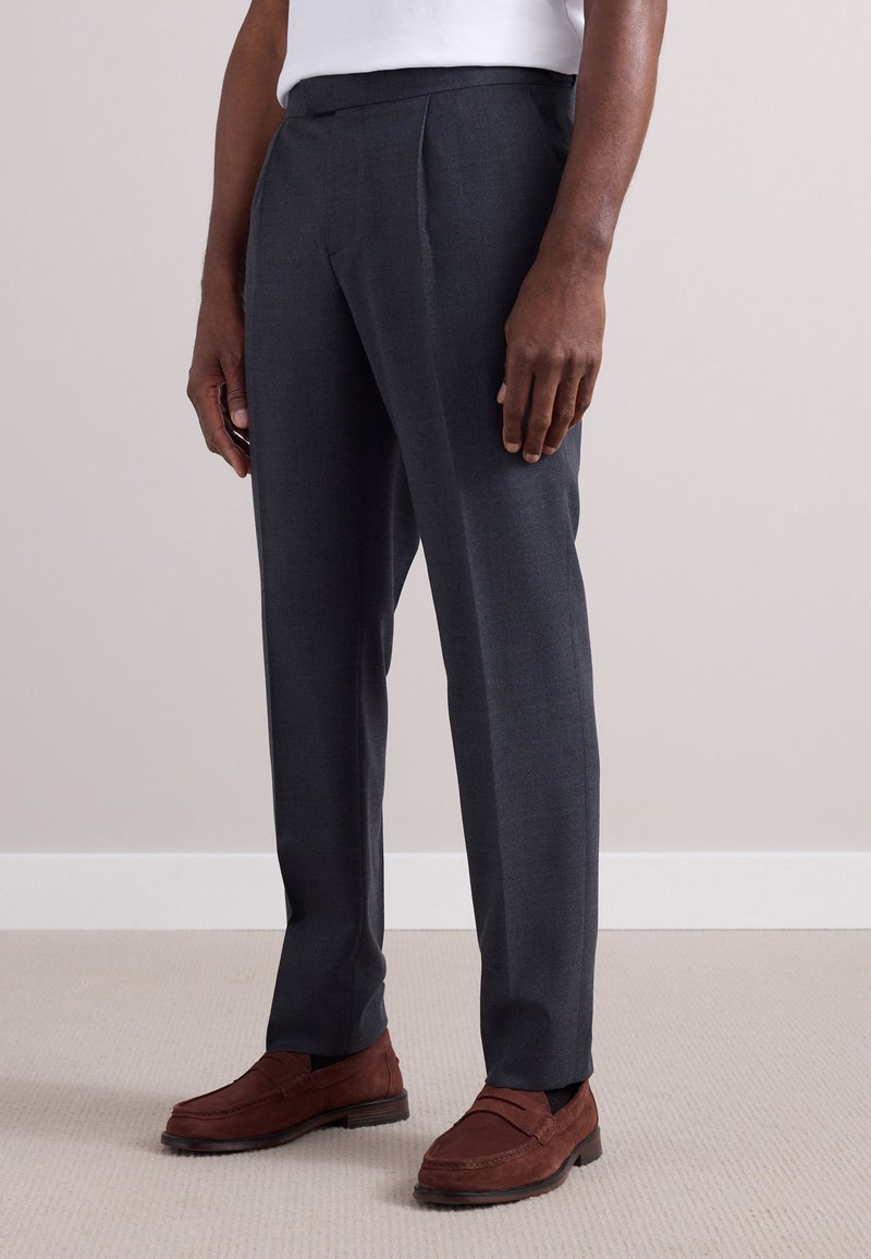 Gray tailored trousers with a flat front, featuring a slim fit and crease detailing. Paired with brown suede loafers on a textured floor.