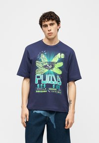 Νεαρός με κατσαρά μαλλιά που φοράει μια μπλε ναυτική φαρδιά μπλούζα με μεγάλο πράσινο λουλούδι και το λογότυπο "PUMA".