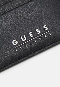 Portafoglio per carte in pelle sintetica nera con superficie texturizzata, logo "GUESS" in rilievo e anno di fondazione, con dettagli di cucitura.