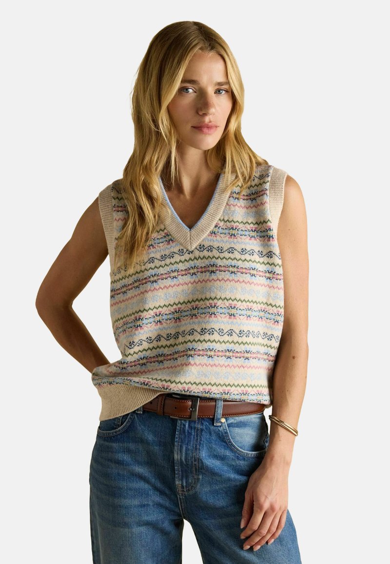 Frau trägt einen ärmellosen Strickpullover mit pastellfarbenen Mehrfarbmuster, blaue Jeans, einen braunen Gürtel und goldene Armbänder am Handgelenk.