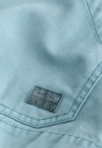Tissu bleu clair avec une petite étiquette rectangulaire cousue, comportant un texte et un logo de couronne près de la couture.