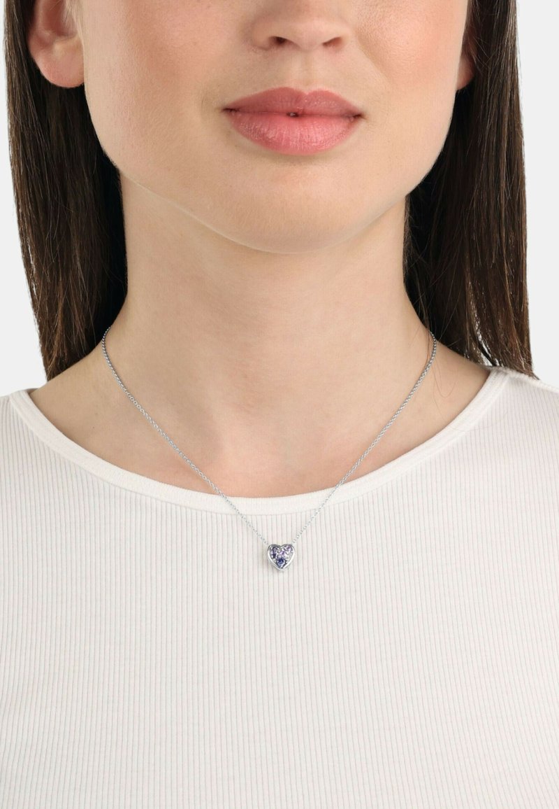 Collana con pendente a forma di cuore in argento, con una pietra centrale viola, indossata su una maglietta chiara.
