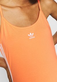 Orange tanktop tillverkad av mjukt tyg, med rund halsringning och tunna axelband. Visad logotyp i vitt i mitten och vita sidränder.