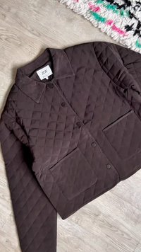 Σκούρο καφέ καπιτονέ μπουφάν με γιακά, κουμπιά τύπου σναπ, τσέπες με καπάκι και μακριά μανίκια, τοποθετημένο σε ανοιχτόχρωμο ξύλινο πάτωμα κοντά σε πολύχρωμο χαλί.