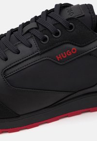 Scarpa sportiva nera con una parte superiore texturizzata, lacci neri, dettagli rossi e una suola rossa, con un logo ben in evidenza.