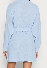 Světle modrý pletený cardigan s páskem v pase, oversized rukávy a texturovaným vzorem. Spodní lem končí nad polovinou stehenní části.