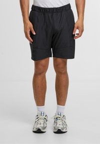 Sorte treningsshorts med elastisk midje, sidelommer og en glatt tekstur. Brukt med hvite ankelsokker og sølvfargede joggesko.