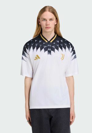 Persona joven con cabello rubio largo que lleva una camiseta de fútbol con estampado geométrico en blanco y negro, con logos dorados y pantalones negros.