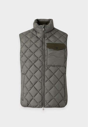 Homme portant une veste matelassée verte sur un pull beige en maille, jean foncé et baskets beiges, debout avec une main dans la poche.