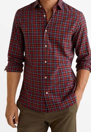 Homme portant une chemise à manches longues à carreaux rouges et bleus, manches retroussées, associée à un pantalon vert olive.