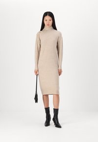Abito lavorato a maglia beige con collo alto, maniche lunghe e tre bottoni decorativi su una spalla; abbinato a stivaletti neri alla caviglia e una borsa piccola.