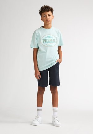 Lichtblauw katoenen T-shirt met "PETROL"-opdruk, gecombineerd met marineblauwe korte broek. Witte sneakers en patroon sokken maken de outfit compleet.