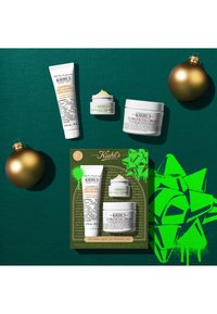 Ensemble de soins Kiehl's comprenant un nettoyant visage à la calendula, un traitement crémeux pour les yeux, une crème ultra visage sur fond vert avec des ornements de Noël dorés.