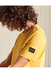Camiseta de algodón amarilla de manga corta, con un parche de marca negro en la manga izquierda y un gráfico sencillo en el frente.