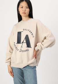 Felpa beige chiaro con vestibilità over size, caratterizzata da un design grafico blu navy che recita "LA California Pacific Dreams" sul davanti.