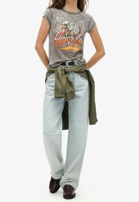 Šedé grafické tričko s designem orla a nápisem "Superdry", v kombinaci se světle modrými širokými džíny a zelenou bundou uvázanou v pase.