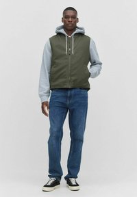 Grön quiltad väst över en grå hoodie, med dragkedja framtill och en känguruficka. Bärs med blå jeans och svarta sneakers.