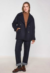 Marinepeacoat met dubbelzijdige knopen, grote revers en zijzakken. Gecombineerd met een bruin gebreid sweater en donkere spijkerbroeken.