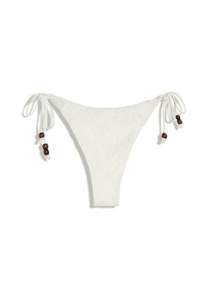 Jonge vrouw in een crèmekleurige bikini en gehaakte hoed staand voor een rotsachtige achtergrond, kijkt recht in de camera.
