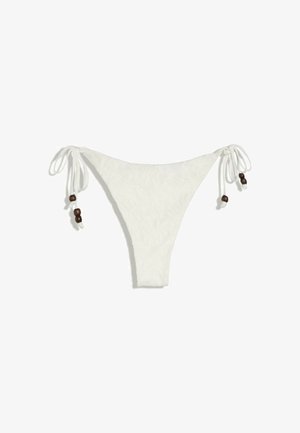 Jeune femme en bikini crème et chapeau en crochet, se tenant devant un arrière-plan rocheux, regardant directement la caméra.