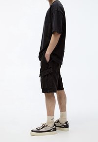 T-shirt noire oversize avec détails usés, associée à un short cargo noir avec poches latérales. Porte des chaussettes beiges et des baskets noires.