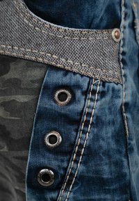 Vêtement en denim avec une ceinture tissée grise, des boutons-pression en métal et un tissu camouflage. Tons de bleu avec des détails de couture contrastants.