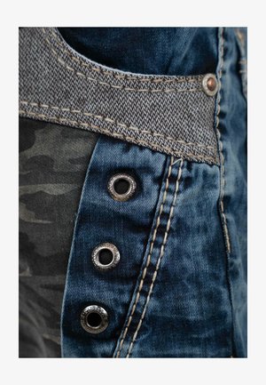 Denim kledingstuk met een grijs geweven tailleband, metalen drukknoopjes en camouflage stof. Blauwe tinten met contrasterende stikseldetails.