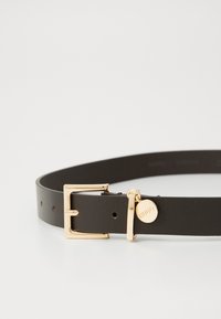 Cintura in pelle marrone con una fibbia in metallo dorato e un piccolo ciondolo. Texture liscia, design classico e una finitura pulita con più fori di regolazione.