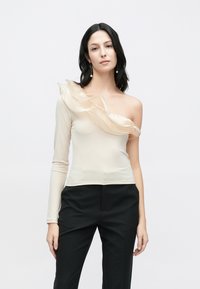 Beige enaxlad topp med en flödande volangdetalj, lång ärm på ena sidan, slät textur, kombinerad med svarta byxor.