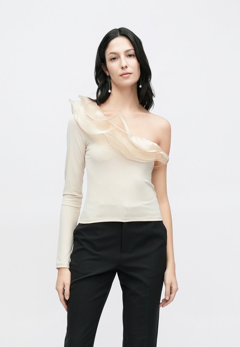 Smėlio spalvos vieno peties palaidinė su lengvai krentančia volaninės detalės, viena ilgomu late, lygiu tekstūra, derinama su juodomis kelnėmis.