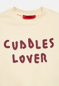Kreminės spalvos džemperis su bordo spalvos užrašu „Cuddles Lover“ ir raudonu prekės ženklo etikete ant apykaklės viduje.