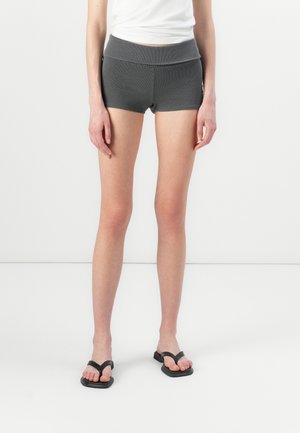 Shorts gris à côtes avec une large ceinture, présentant un design ajusté. Portés avec des tongs noires, affichant un look simple et décontracté.
