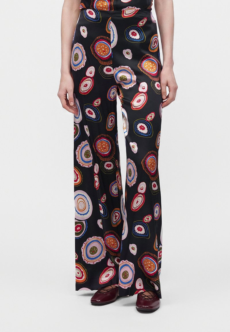 Pantalon noir à jambes larges avec des motifs circulaires colorés, porté avec des chaussures plates rouge foncé, montré de la taille aux pieds.