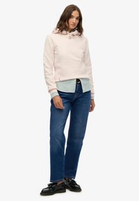 Sudadera rosa claro con cordón, bolsillo canguro y puños acanalados, superpuesta a una camisa a rayas. Combinada con jeans azules y zapatos negros.