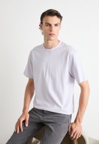 T-shirt violet clair à manches courtes en tissu doux, avec un col rond et un logo en relief discret à l'avant. Associé à un jean gris.