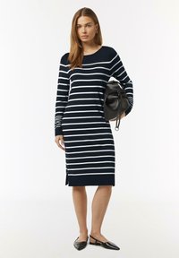 Marineblaues Kleid mit weißen horizontalen Streifen, langen Ärmeln, seitlichem Schlitz und geripptem Kragen. Das Modell hält eine schwarze Tasche und trägt flache schwarze Schuhe.