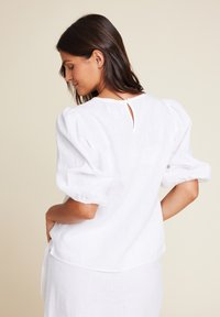 Hvit bluse med puff-ermer laget av lin, med rund halslinning og hjerteformet detalj bak. Uformelt design, litt løs passform.