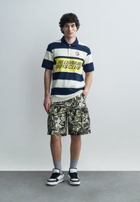 Junger Mann steht, trägt ein marineblau-weiß gestreiftes Polo mit der Aufschrift "Billionaire Boys Club", Tarn-Cargoshorts, graue Socken und schwarz-weiße Sneaker.