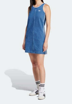 Femme portant une robe en jean bleue sans manches avec des chaussettes blanches à rayures et des baskets blanches à rayures noires.