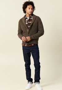 Cardigan marrone a maglia con colletto a scialle, chiusura con bottoni, indossato su una camicia a quadri. Jeans scuri e sneakers bianche completano l'outfit.